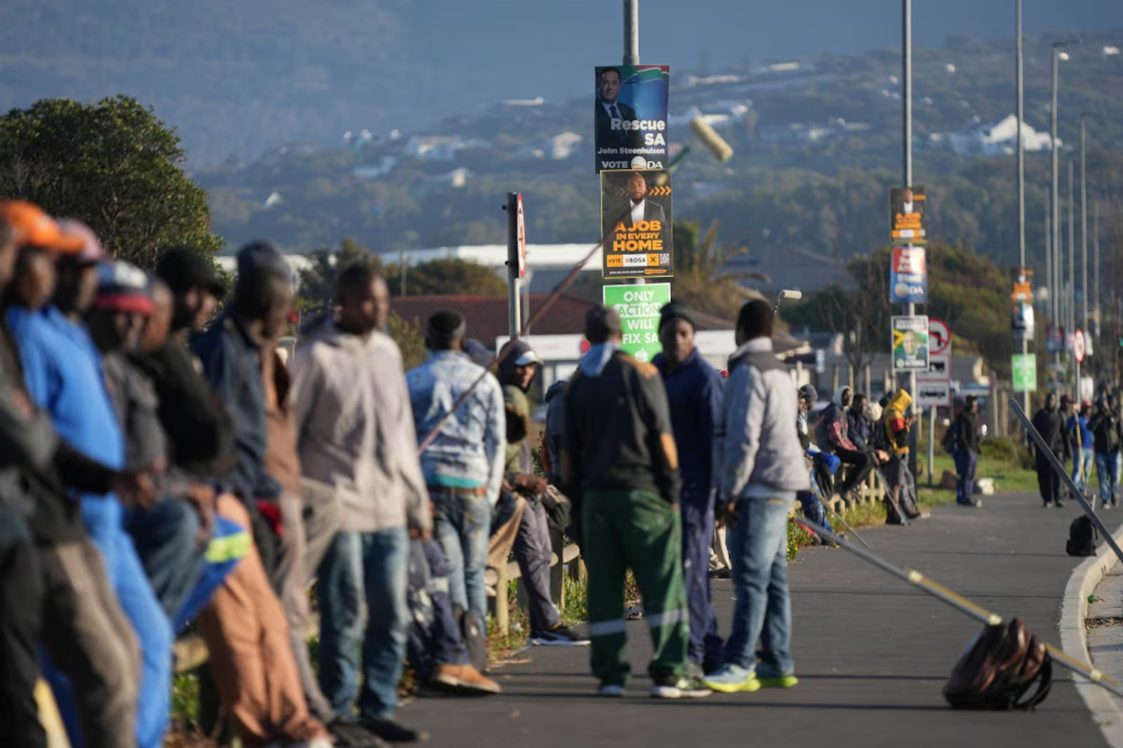 Unemployed job seekers line a street waiting for casual employment, as they sit beneath election campaign posters for the South African general elections